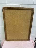 Photo of a vertically oriented rectangular cork board with rope trim on the wood frame, showing texture and condition.