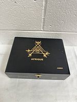 Closed black wooden cigar box with Monte Cristo Afrique gold emblem on top and Jambo text on corner.