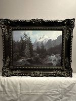 Frontal photo of framed landscape painting showing mountain, trees and river with cloudy sky.