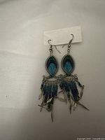 Front view of pair of boho style earrings showing beaded oval centerpiece with feather design, blue and green beads, and dangling bead clusters with metal feather charms.