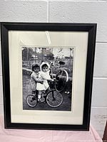 Frontal image showing black photo frame with black and white photo of two children on tricycle outdoors.