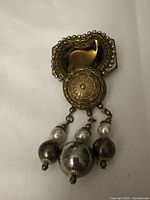 Metal runework brooch with central circular motif and three dangling beads in silver and pearl tones, shown on a plain neutral background.