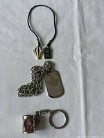Three pieces of costume jewellery laid out on a white background, showing a blue fabric necklace with a hot air balloon pendant and rectangular tag, a silver-tone chain necklace with a maple leaf dog tag, and a metal keychain.