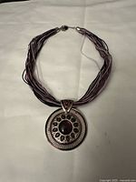 Full view of the silver tone metal pendant necklace showing the entire pendant and multi-strand purple cord on light background