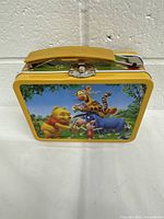 Front view of tin lunch box showing colorful Pooh and friends decor with yellow handle and metal latch.