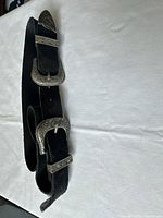 Black suede belt displayed coiled with visible silver engraved buckles and loops, showing belt's material and metal detailing