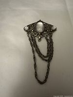 Front view of vintage brooch showing central pearl-like stone, side stones, intricate filigree metal work and hanging chains.