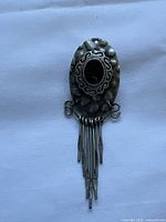 Front view of an oval vintage brooch made of silver colored metal with a black central stone and attached metal tassels.