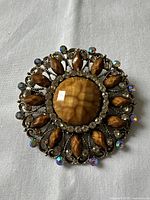 Close-up image of the vintage brooch showing central large brown-yellow faceted stone, surrounding marquise brown stones, clear rhinestone circle, and multicolored rhinestone accents on dark metal setting.