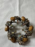 Brooch laid flat on a light fabric background showing top view with leaf-shaped brown accents and several multicolored rhinestones.