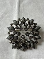 Front view of vintage circular brooch showing gray-black faceted marquise stones and round rhinestones in an ornate leaf-wreath design.