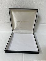Front view of black rectangular vintage jewelry display gift box opened to show cream satin-like lid lining with gold embossed logo and white cushioned base insert.