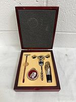 Open wooden box showing wine accessory set including corkscrew, stopper, pourer, drip ring, and thermometer in fitted insert.