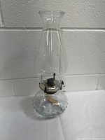Front view of the clear glass oil lamp showing the base, metal burner, and tall chimney against a gray wall.