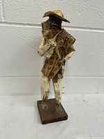 Full view of orange-brown papier mache man figurine with poncho, hat, and sack on a wood base against cinderblock wall.