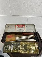 Top view of open metal case showing cleaning rods, brushes, plastic pouches, and printed instruction label inside lid with Outers branding and use instructions.