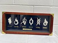 Front view of vintage framed sailor's knots display showing six distinct white rope knots with labeled gold-tone plates against black background in wood frame with brass corners.