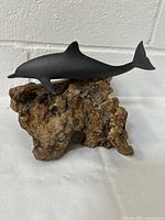 Gray dolphin sculpture mounted on a knotty, natural driftwood base with visible texture and shape variations. Small medallion plaque present on driftwood.