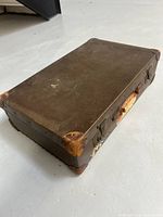 Full view of rectangular brown vintage Vulcanfibre suitcase with scuffed leather corners and metal latches, closed and viewed from an angle on a white floor.