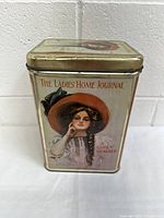 Front view of the vintage metal tin box showing a young woman in a brown hat, titled The Ladies Home Journal Girls' Number.