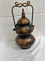 Front view of vintage bronze urn with handle, lid attached by chain, and grape leaf decorations.