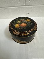Side angle showing the round black tin with floral lid, gold scroll detailing, and slight patina.