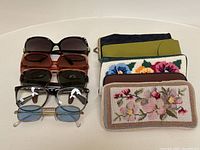 Six eyeglasses and eyeglass cases arranged, including black sunglasses and five assorted cases with floral needlepoint and other designs.