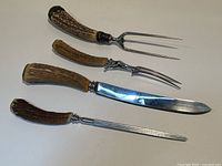Four carving utensils laid out on white surface. Each has an antler handle with silver mounts and a metal blade or fork tines. The sizes and shapes vary including fork, knives and sharpening steel.