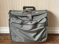 Front view of green grey Briggs & Riley rolling wardrobe bag showing upright design, handle, and zippered front pocket.