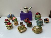 Photo showing all 14 trinket boxes including a watering can, basket with flowers, multiple ladybird topped boxes, heart-shaped butterfly box, clock box, and bee decorated boxes arranged on white surface.