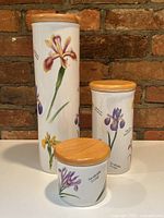 Front view of the set of three white ceramic kitchen canisters with colorful iris flower designs by Sky McGhie on a brick background, wooden lids on top.