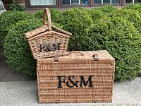 Photo of two wicker picnic baskets placed outside in front of greenery showing the 'F&M' lettering and leather strap closures.