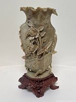 Full view of beige-gray soapstone vase with floral and bird relief carving on a carved dark wooden base