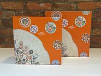 Two orange Hermès square boxes with colorful floral circular patterns on the front against a brick wall background.
