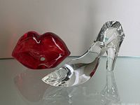 Red glass lips sculpture alongside clear crystal shoe figurine on a reflective surface