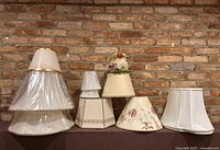 All lamp shades arranged in a row against brick wall.