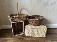 Lot of three woven baskets placed on wooden floor against a beige wall. Includes a tall rectangular handled basket, a round basket, and a rectangular lidded basket seen from front.
