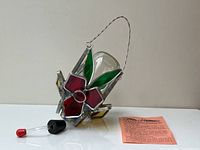 Front and angled view of stained glass hummingbird feeder with red, green, and yellow glass petals/leaves, clear glass jar inside frame, metal hanging wire, and small glass dropper with rubber stopper lying in front beside orange instructions sheet.
