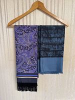 Two vintage men's scarves hanging on a wooden hanger against a striped wallpaper background. Left scarf is purple and navy paisley with black fringe. Right scarf is black and slate blue with subtle textured pattern and short fringe.
