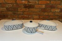 Front view of three Lyngby Denmark ceramic cookware pieces on white surface with brick wall background. Two rectangular roasting dishes with lids flank a round saucepan with lid and handle in center.