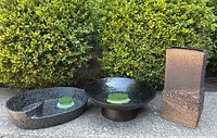 Three metal or metallic pottery flower vessels outdoors by green shrub. Oval vessel has a flower frog divider, round pedestal bowl has a flower frog, rectangular vase has textured finish.