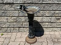 Front view of full cast metal bird bath showing bowl, pedestal, and base against masonry wall