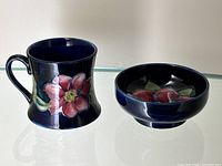 Miniature cobalt blue Moorcroft mug with clematis flower pattern and matching dish also with clematis decoration, displayed on glass surface.