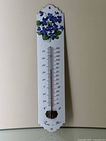 Full shot of the metal wall-mounted thermometer with porcelain enamel surface showing floral artwork on top and Panchet à Paris label at bottom.