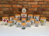 Full group of 13 porcelain canisters with botanical designs and bamboo lids displayed against brick wall
