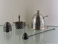 Photo of all four silver plated bee hive themed items on glass surface: salt shaker, basket, honey pot with lid and spoon, candle snuffer with beehive shaped snuffer head