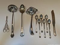 Overview of all 10 silver plated serving utensils on white background showing a pair of scissors, ladle, large serving spoon, strainer spoon, six teaspoons, and serrated knife with some tarnishing visible.