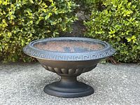 Front angle of small dark cast iron oval planter on pedestal base showing decorative rim and scalloped body detail.