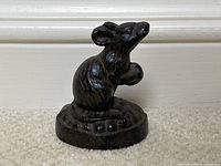 Front view of small wrought iron mouse door stopper on carpeted floor against white baseboard.