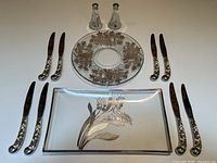 Eight cutting knives arranged neatly around two glass plates with silver overlay designs, showing the detailed floral patterns on the utensil handles and plates.
